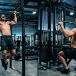 Deux hommes musclés s'entraînent aux barres parallèles en salle.